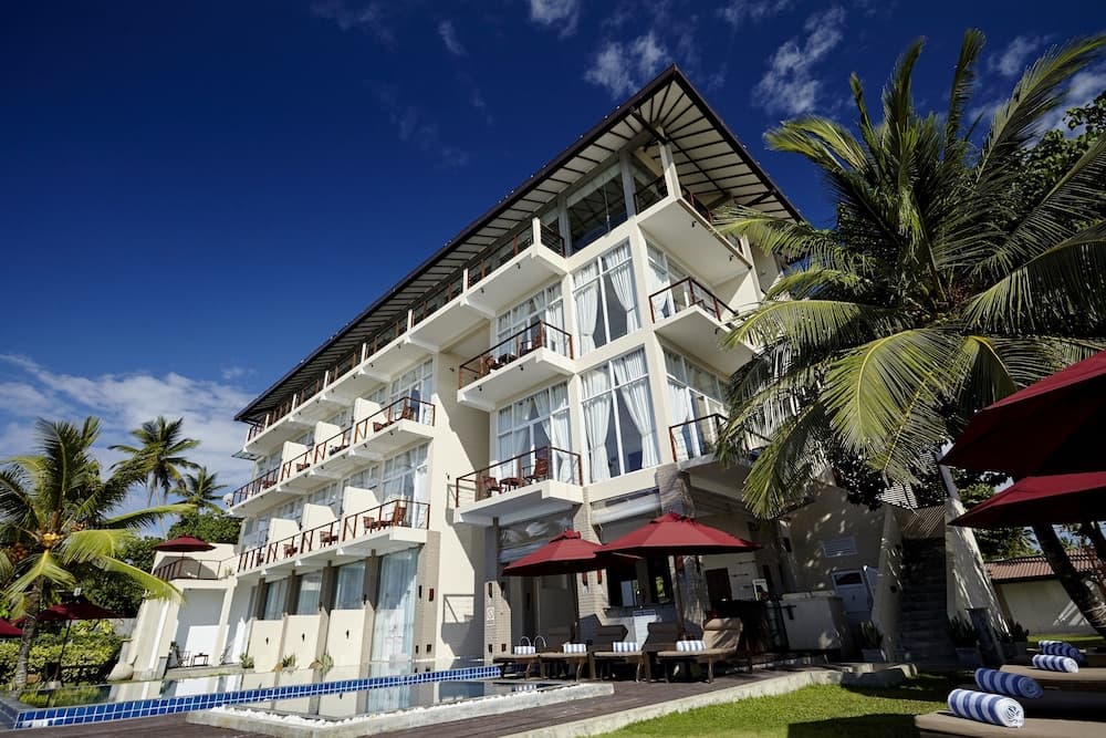 A serene tropical resort features a multi-story building with balconies under a clear blue sky, surrounded by lush palm trees, vibrant red umbrellas, and inviting lounge chairs, perfect for a relaxing vacation getaway.