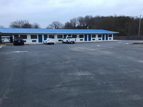 A quiet roadside motel with a blue roof and cracked parking lot exudes a nostalgic charm, ideal for travelers seeking budget-friendly accommodations in a serene suburban setting.