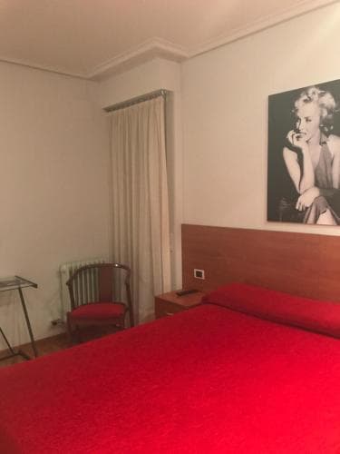 A cozy hotel room features a vibrant red bedspread, a wooden headboard, a Marilyn Monroe poster, and simple furnishings, creating an intimate and nostalgic atmosphere ideal for travelers seeking a charming stay.