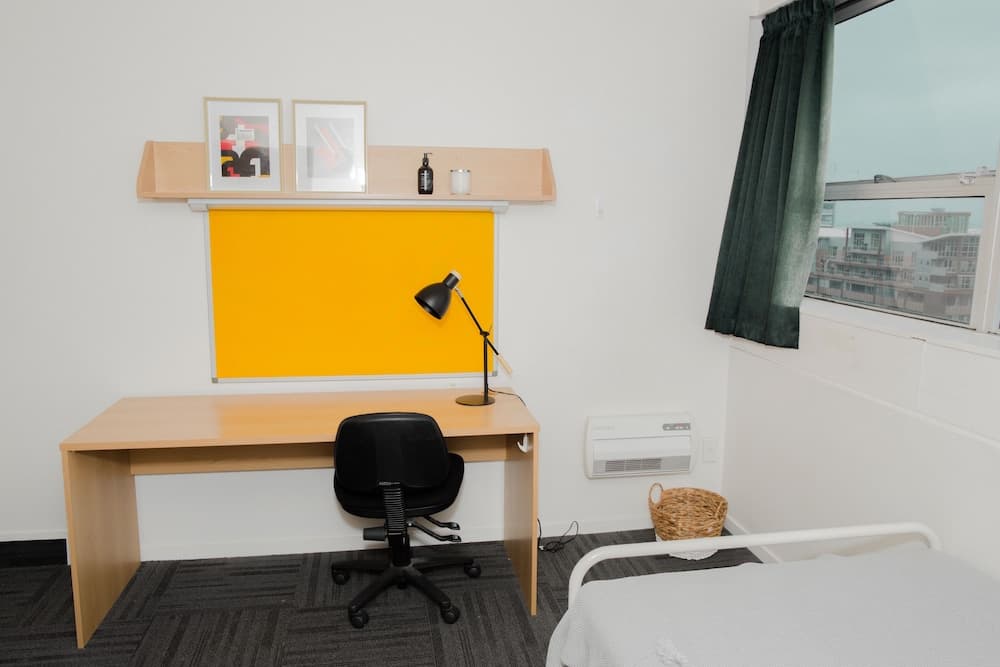 A cozy dorm room features a wooden desk with a black chair, a vibrant yellow bulletin board, framed artwork, and a window view of urban buildings, ideal for students seeking a functional and serene study space.