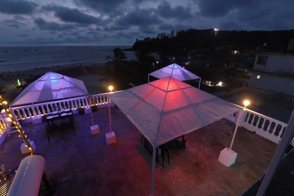 An atmospheric evening view of a rooftop terrace adorned with illuminated tents and string lights, overlooking a serene beach under a moody sky, perfect for romantic dinners or intimate gatherings.