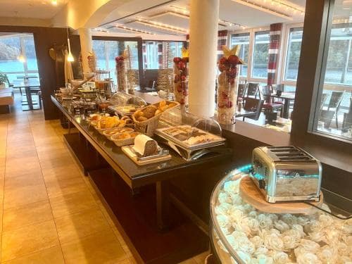A cozy hotel buffet area with a variety of breakfast items, a vintage toaster, and large windows offering a serene lakeside view, perfect for travelers seeking a relaxing morning start.