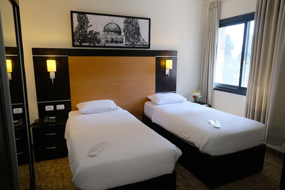 A cozy hotel room features two neatly made twin beds under warm lighting, framed by a scenic artwork above the wooden headboard, ideal for travelers seeking comfort and tranquility.