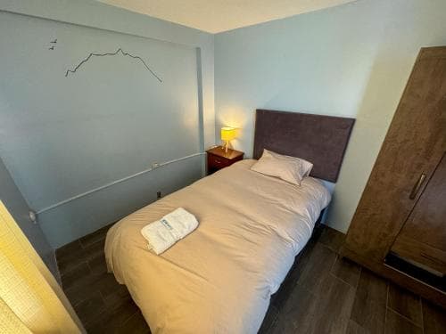 A cozy hotel room features a neatly made bed with soft lighting, minimalist decor including a sketched mountain on the wall, and wooden furnishings, ideal for travelers seeking a simple and restful stay.