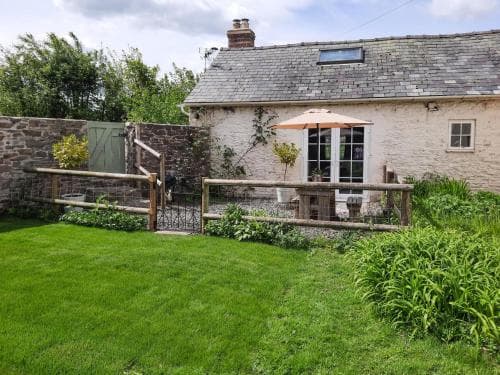 A charming countryside cottage with a lush green lawn, wooden fence, and patio area under an umbrella invites travelers seeking a peaceful rural retreat.