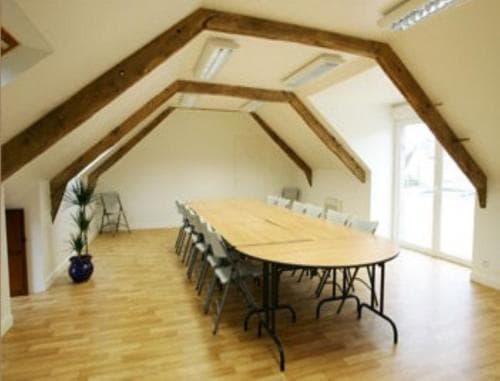 A cozy attic meeting room with exposed wooden beams, a large oval table surrounded by chairs, and natural light streaming through windows, ideal for intimate business gatherings or retreats.