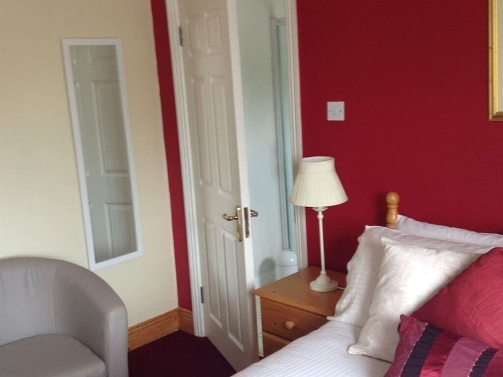A cozy bedroom with vibrant red walls, a neatly made bed adorned with white and red pillows, a wooden bedside table with a lamp, and a comfortable armchair, ideal for travelers seeking a warm and inviting accommodation.