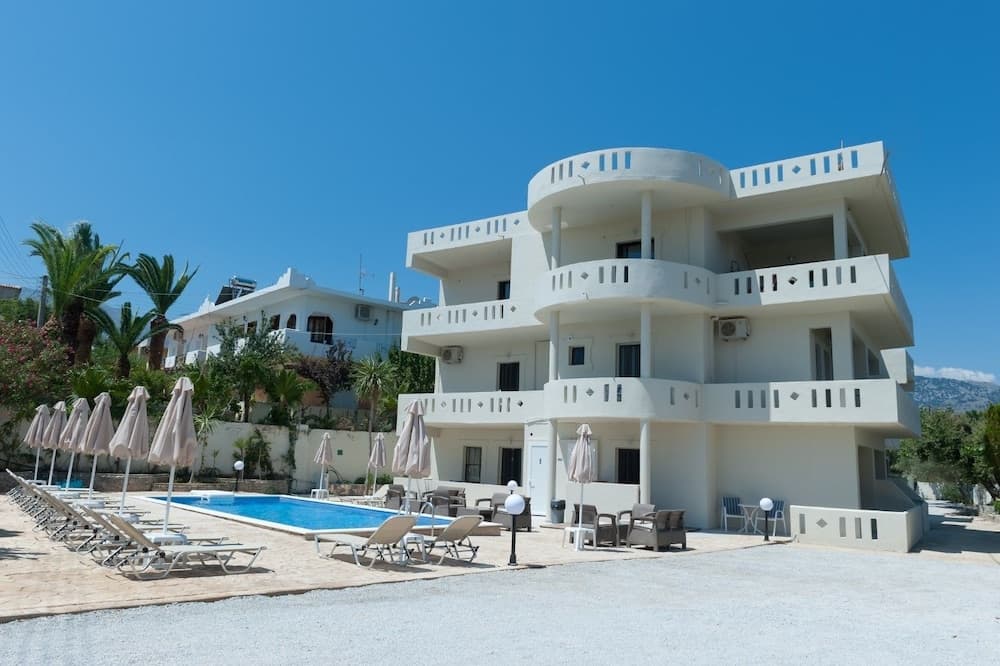 A serene coastal resort features a modern white building with balconies overlooking a tranquil pool area adorned with lounge chairs and umbrellas, perfect for a relaxing vacation getaway.