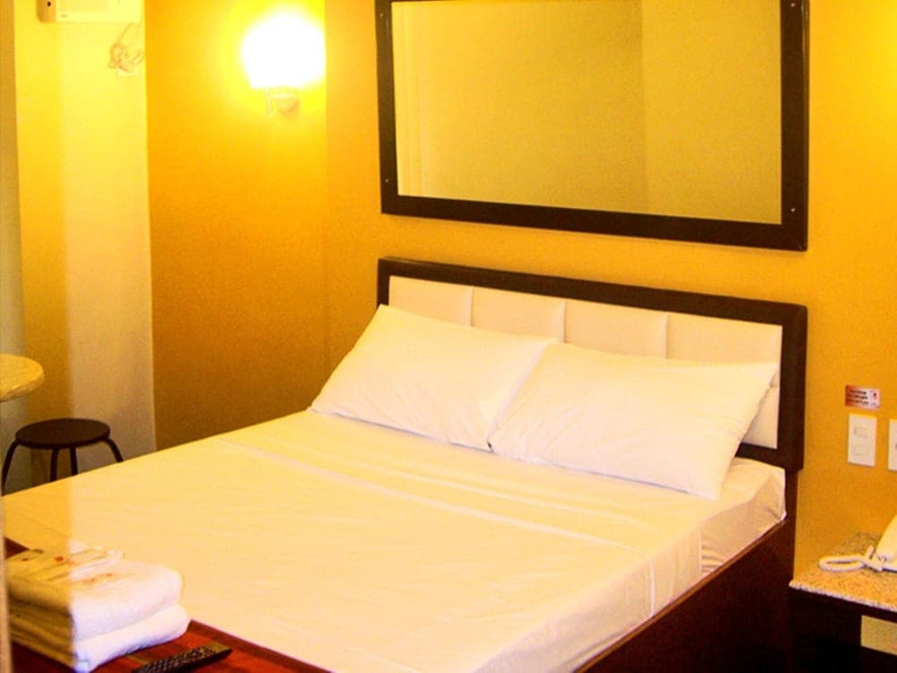 A cozy hotel room with warm yellow walls, a neatly made bed with white linens, a wall-mounted mirror, and bedside amenities creates an inviting atmosphere for travelers seeking comfort.