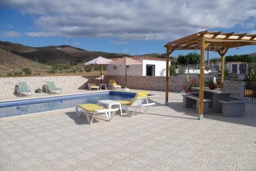 A serene outdoor villa scene features a tiled patio with a vibrant blue pool, lounge chairs, a wooden pergola, and distant hills under a partly cloudy sky, ideal for a relaxing vacation getaway.