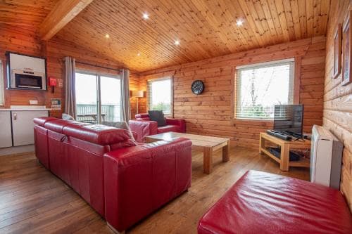 A cozy cabin interior with warm wooden walls and ceilings, vibrant red leather sofas, and natural light streaming through large windows, ideal for a relaxing getaway or family vacation rental.