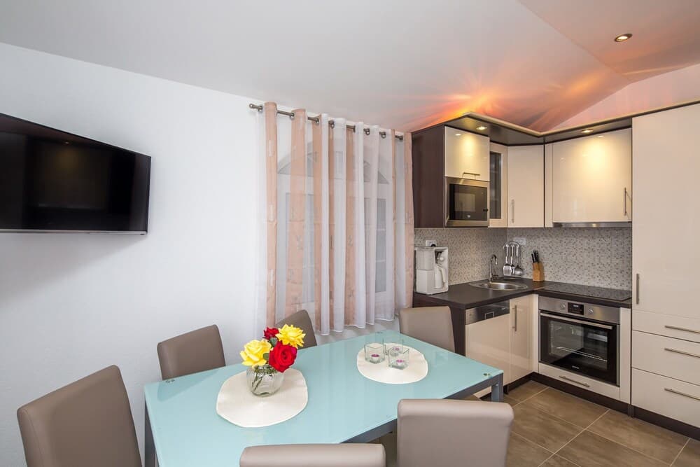 A cozy apartment kitchenette features a modern dining area with a blue table adorned by vibrant flowers, sleek white cabinetry, and warm lighting, ideal for travelers seeking a comfortable self-catering stay.