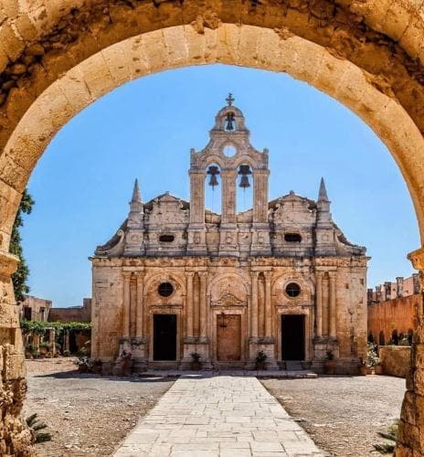 A serene courtyard framed by an ancient stone archway leads to a majestic, weathered church under a clear blue sky, ideal for cultural heritage exploration and photography.