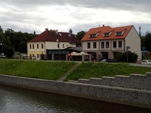 A serene riverside scene featuring quaint buildings with red-tiled roofs, outdoor seating under umbrellas, and a stone embankment, ideal for a peaceful getaway or a leisurely stroll along the water.