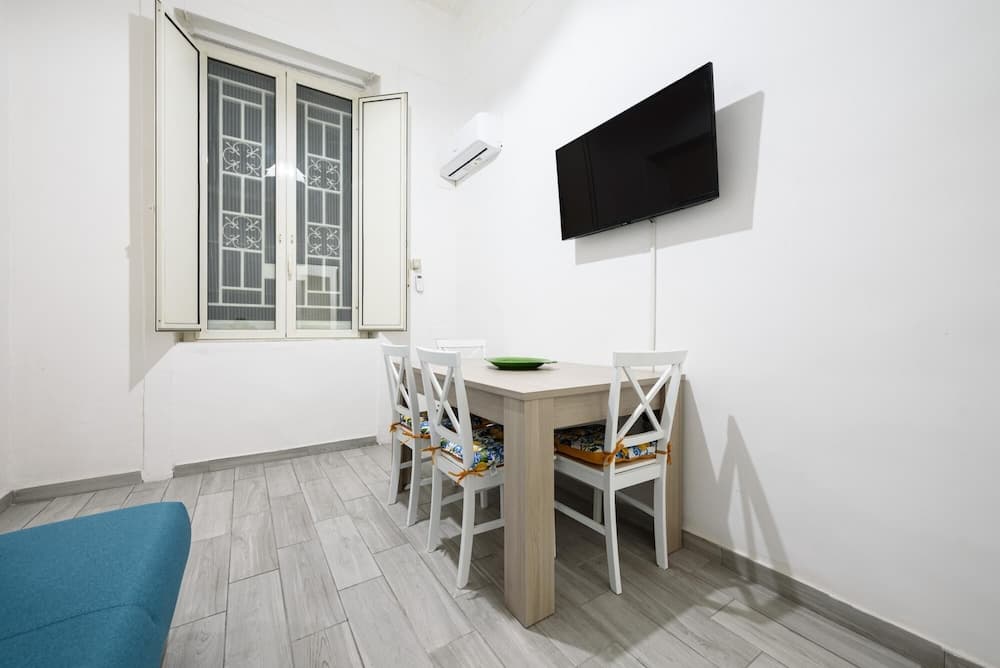 A cozy apartment dining area features a wooden table with white chairs adorned with colorful cushions, a wall-mounted TV, an air conditioner, and a window with decorative shutters, ideal for travelers seeking comfortable and modern accommodations.