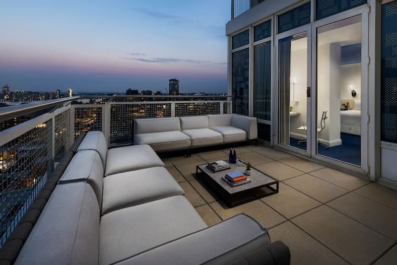 A serene rooftop terrace at dusk in New York City features modern white sofas, a coffee table with books, and a view of the illuminated skyline, ideal for luxury urban retreats.