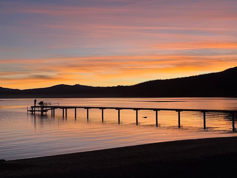 A serene lakeside scene at sunset features a long wooden pier extending into calm waters, with silhouetted figures enjoying the tranquil atmosphere, ideal for travelers seeking peaceful retreats in natural settings.