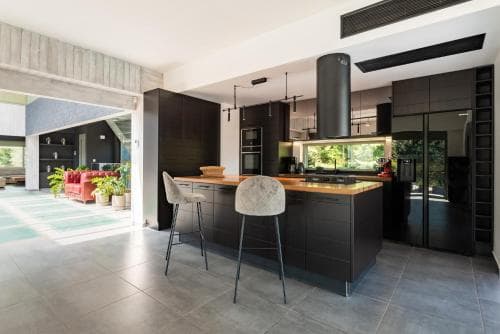 A modern kitchen with sleek black cabinets, a wooden countertop, and bar stools opens to a cozy living area with vibrant red furniture, ideal for travelers seeking stylish and functional accommodations in a contemporary home setting.