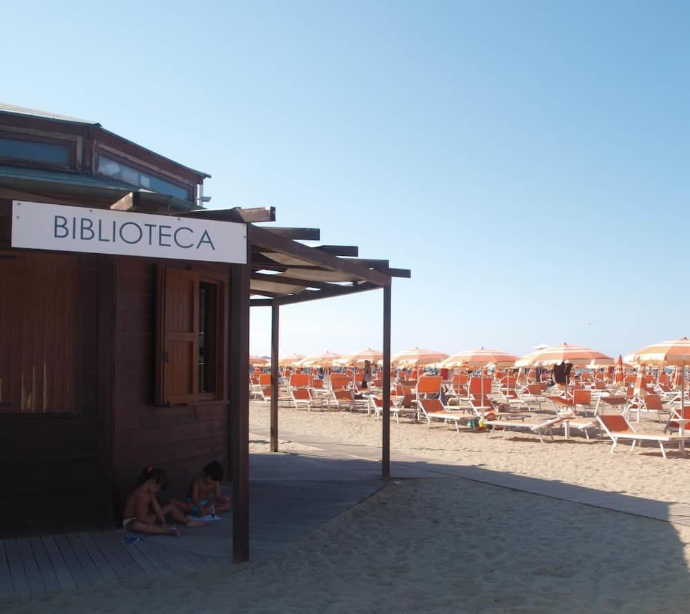 A serene beach scene features a wooden "BIBLIOTECA" hut where children read under the shade, surrounded by rows of orange umbrellas and loungers, ideal for a relaxing family vacation.