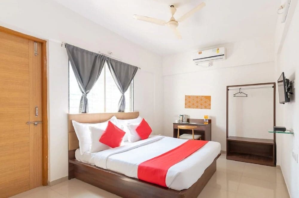 A cozy hotel room features a neatly made bed with red accents, a wooden door, a window with gray curtains, and modern amenities like an air conditioner and flat-screen TV, ideal for travelers seeking comfort and convenience.