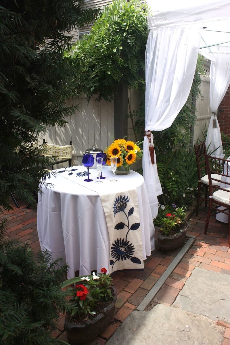 A serene outdoor patio at a boutique guest house features a charming table set with sunflowers, blue glassware, and white drapery, surrounded by lush greenery, ideal for travelers seeking a cozy and intimate dining experience.