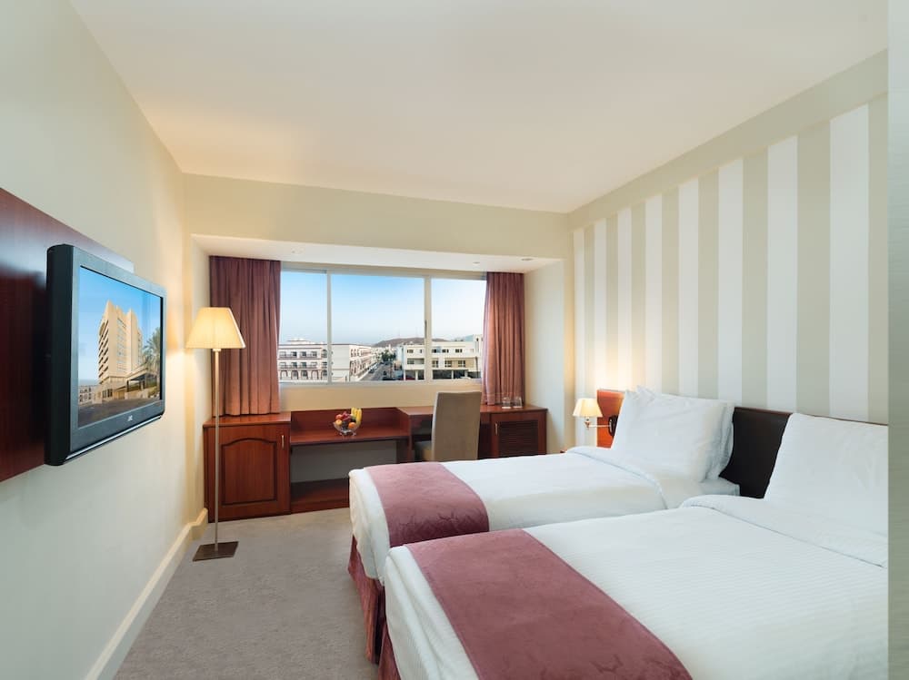 A cozy hotel room with twin beds, striped walls, and a scenic city view through large windows offers a comfortable stay for travelers seeking relaxation and convenience.
