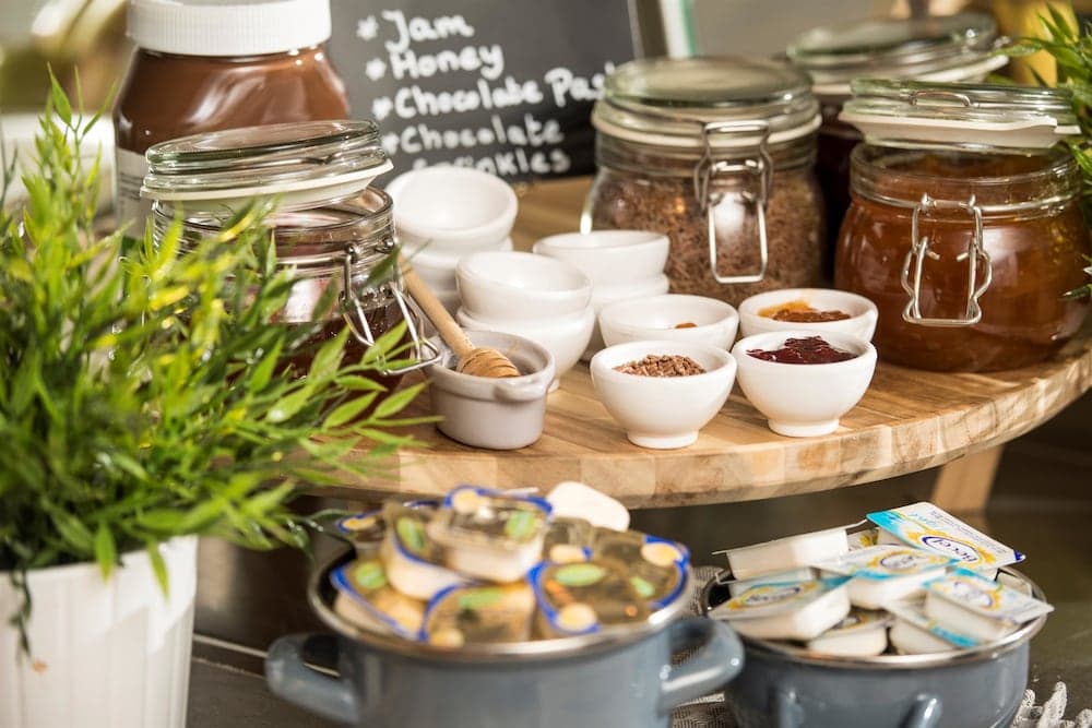 A cozy café setting features a wooden table laden with jars of jams, honey, chocolate pastes, and small bowls of toppings, surrounded by fresh greenery, creating an inviting atmosphere perfect for travelers seeking a delightful breakfast or snack spot.
