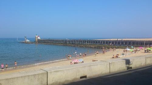 A sunny beach scene with people relaxing under umbrellas and a long pier extending into calm waters, ideal for a leisurely seaside getaway.