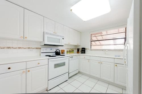 A bright, modern kitchen with white cabinetry, tiled flooring, and natural light filtering through blinds, ideal for travelers seeking a cozy self-catering accommodation.