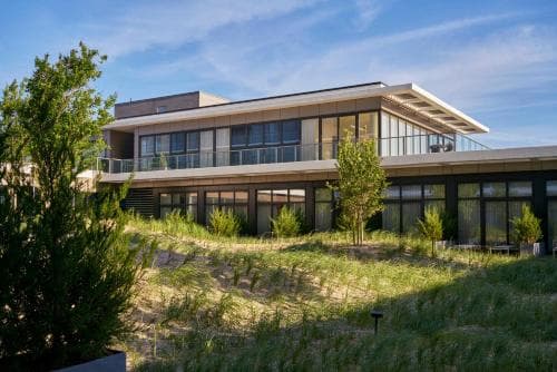 A modern two-story house with large glass windows sits atop a grassy hill under a clear blue sky, offering a serene retreat ideal for eco-conscious travelers seeking luxury and nature.