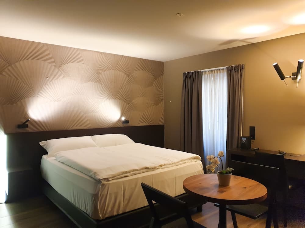 A cozy hotel room features a neatly made bed against a textured wall, soft lighting, a small dining table with chairs, and a window with curtains, ideal for travelers seeking comfort and relaxation.