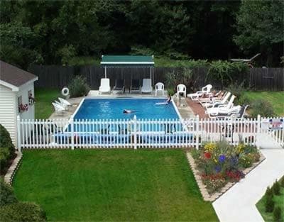 A serene backyard pool area enclosed by a white picket fence features lounge chairs, a green cabana, and lush greenery, ideal for a relaxing family vacation retreat.