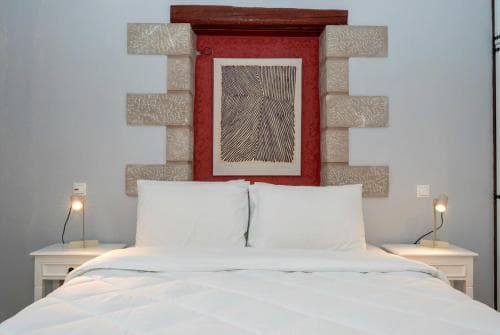 A cozy hotel room features a neatly made bed flanked by white nightstands with warm lamps, set against a minimalist wall adorned with a striking red-framed artwork, ideal for travelers seeking comfort and style.