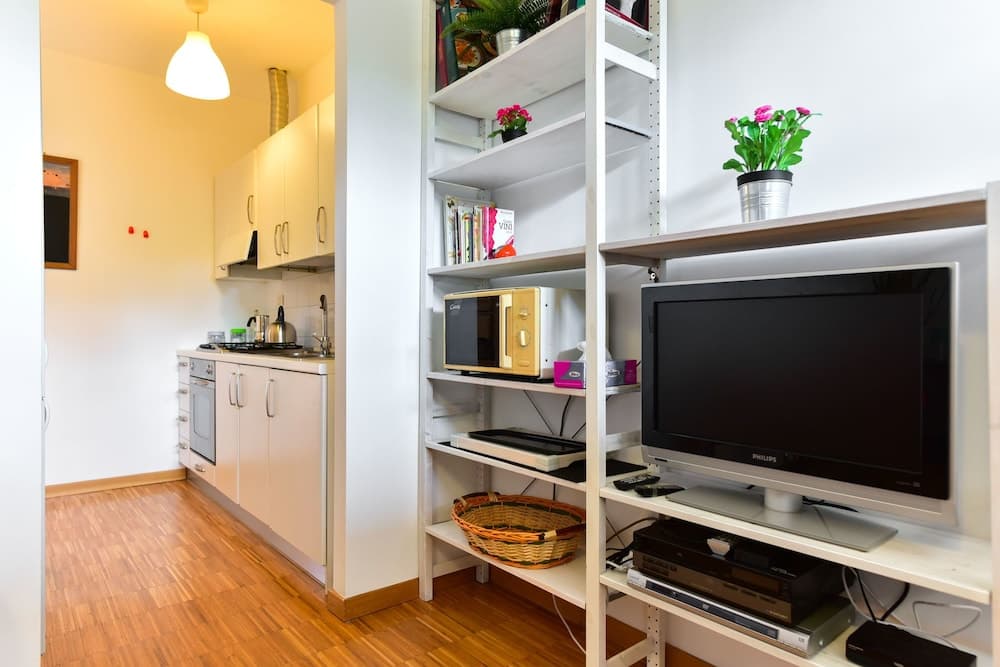 A cozy apartment interior features a compact kitchen with white cabinets and a wooden floor, adjacent to a shelving unit holding a TV, books, plants, and a microwave, ideal for travelers seeking a homey and functional living space.