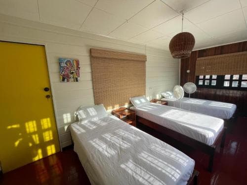 A cozy, sunlit dormitory room with wooden floors, twin beds, a vibrant yellow door, and bamboo blinds evokes a relaxed atmosphere, ideal for budget travelers seeking comfort in shared accommodations.