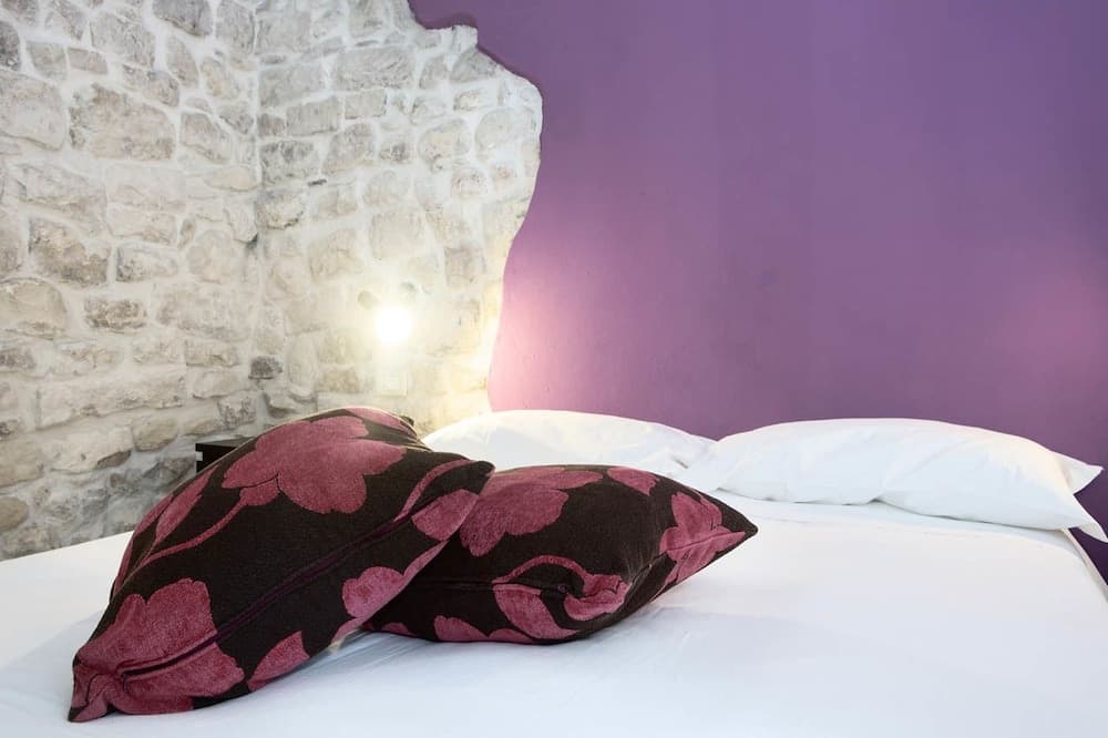 A cozy bedroom with a white bed adorned by floral-patterned pillows, featuring a rustic stone wall and a vibrant purple accent wall, ideal for travelers seeking a charming and intimate accommodation experience.