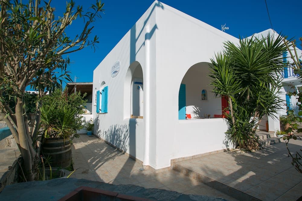 A serene Mediterranean courtyard features a charming white building with blue accents, lush greenery, and tiled pathways, ideal for travelers seeking a peaceful retreat.