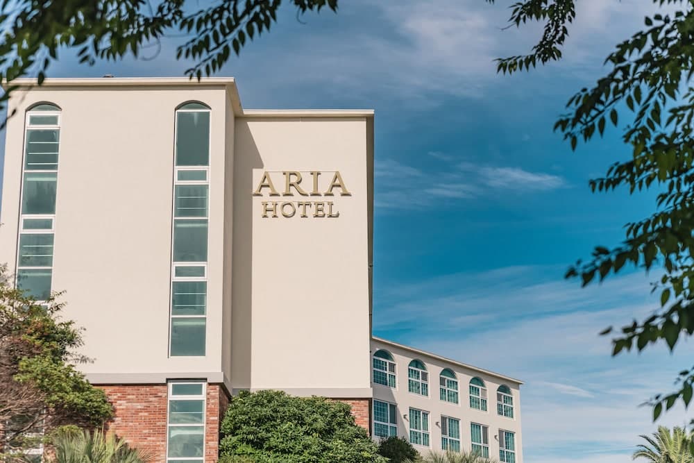 The serene Aria Hotel, framed by lush greenery against a clear blue sky, offers travelers a tranquil urban retreat with its modern architecture and inviting facade.