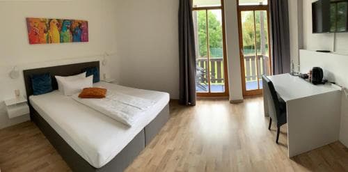 A cozy hotel room with a neatly made bed, wooden flooring, a desk area, and a balcony view of greenery, ideal for travelers seeking comfort and relaxation.