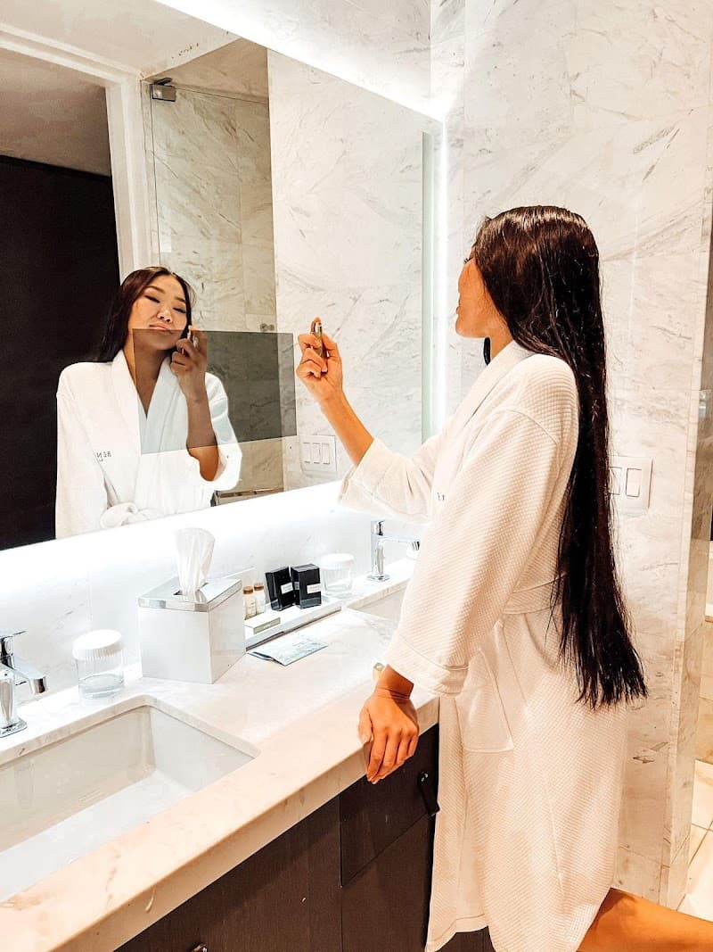 A woman in a white robe applies makeup in the luxurious marble bathroom of a hotel room, creating an intimate and serene atmosphere ideal for travel bloggers showcasing upscale accommodations.