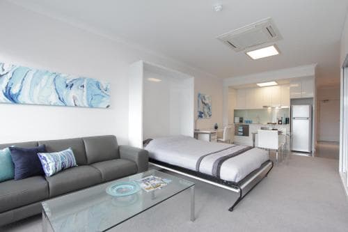A modern studio apartment features an open-plan layout with a cozy sofa, sleek bed, vibrant artwork, and a well-equipped kitchen in the background, ideal for travelers seeking comfort and convenience.