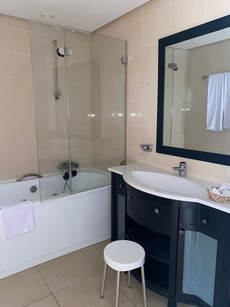 A modern bathroom with beige tiles, a glass-enclosed bathtub, black vanity with a large mirror, and a white stool creates a clean and cozy atmosphere ideal for travelers seeking comfort and functionality.