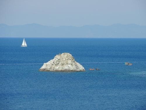 A serene coastal scene features a sailboat gliding across calm blue waters near a rocky outcrop, ideal for travelers seeking tranquil seaside getaways.