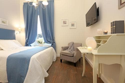 A cozy hotel room features a neatly made bed with blue accents, a wooden desk with a mirror, soft lighting, and a serene ambiance, ideal for travelers seeking comfort and relaxation.