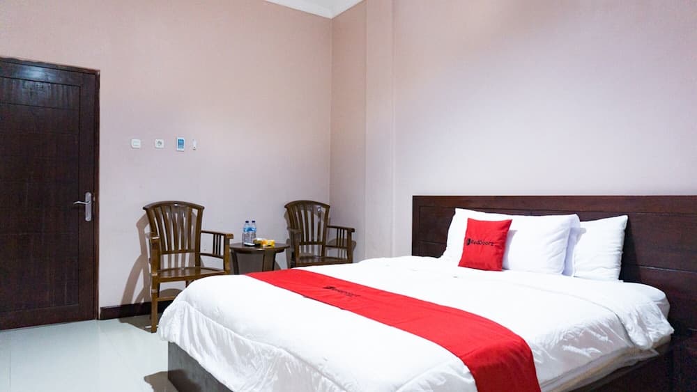 A cozy hotel room with soft pink walls, a neatly made bed adorned with red accents, wooden chairs, and a small table creates a welcoming atmosphere ideal for travelers seeking comfort and simplicity.