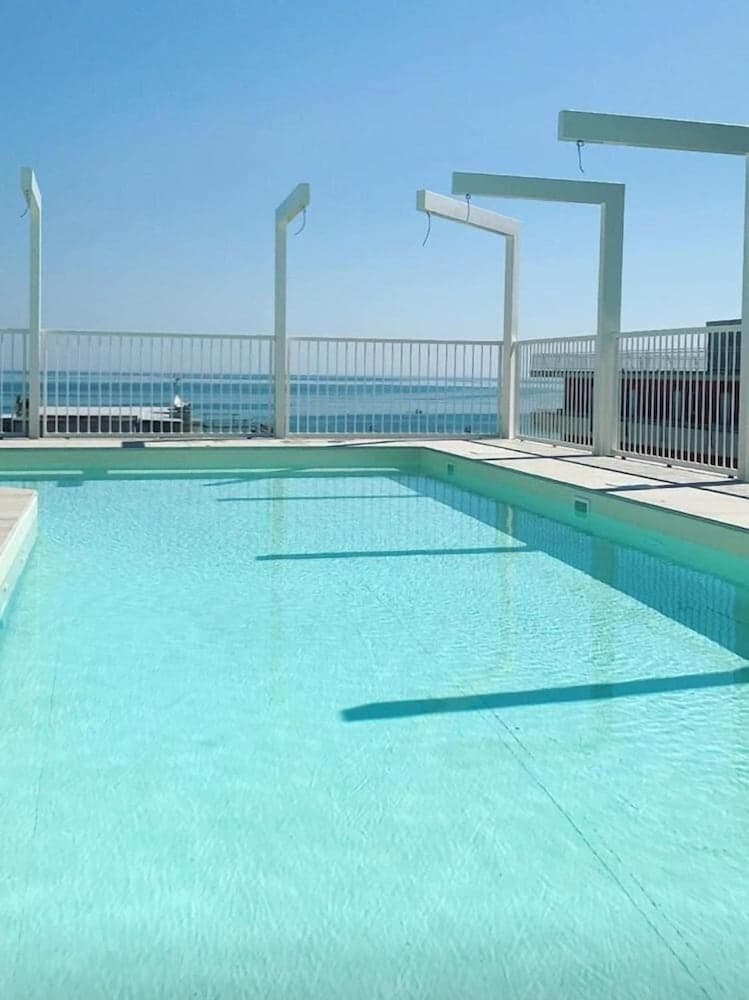 A serene rooftop pool with clear turquoise water overlooks the ocean, framed by white railings and minimalist structures, ideal for travelers seeking relaxation and coastal views.