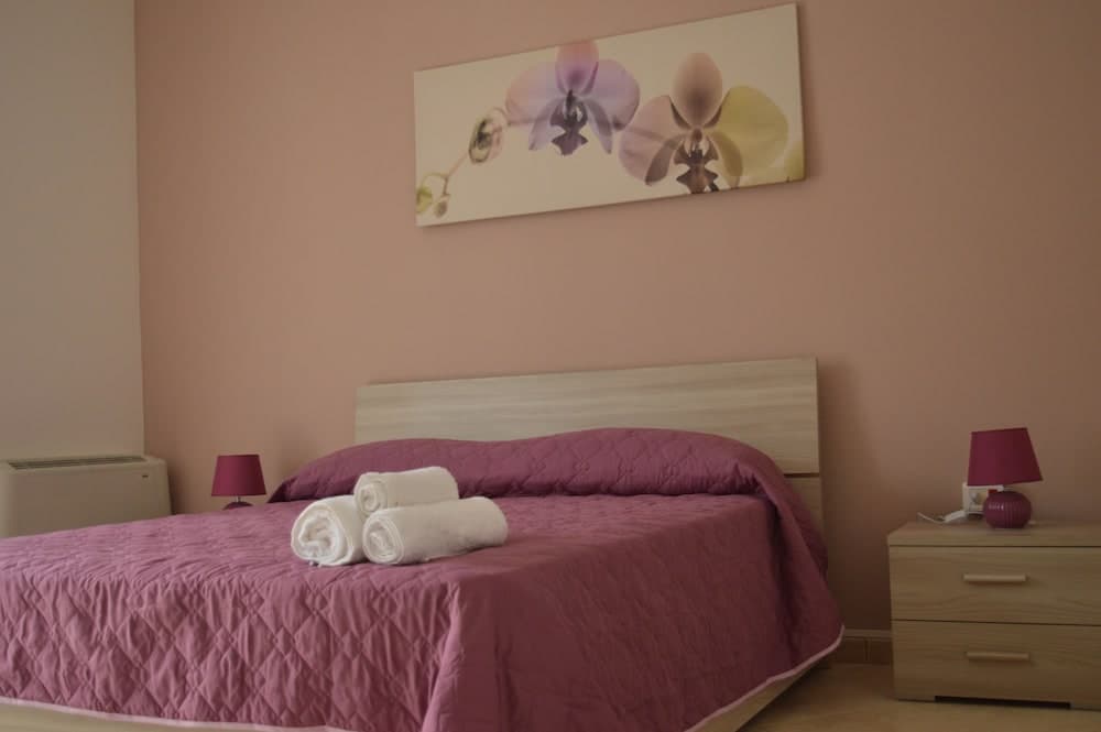 A cozy bedroom with soft pink walls, a neatly made bed adorned with rolled towels, wooden furniture, and floral artwork creates a serene atmosphere ideal for travelers seeking relaxation.