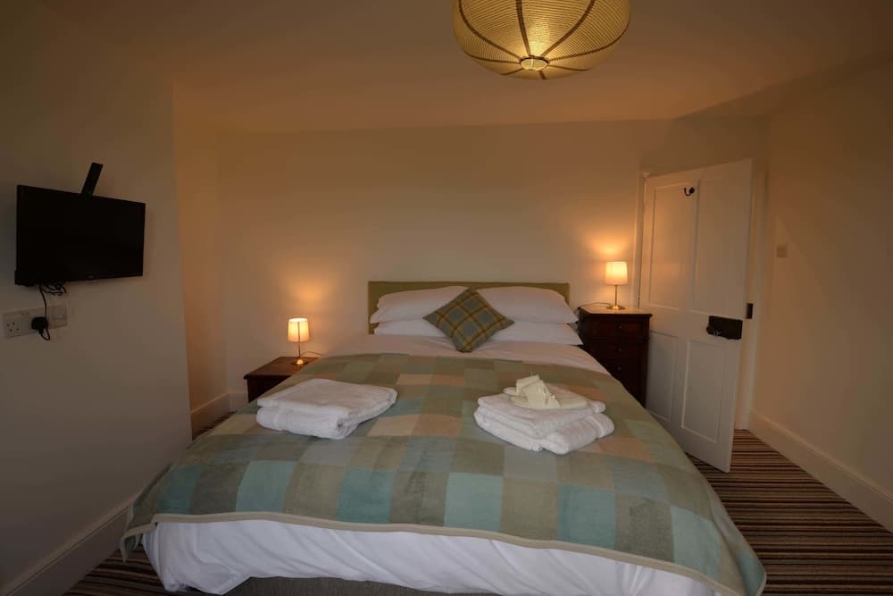 A cozy hotel room features a neatly made bed with folded towels, warm lighting from bedside lamps, a wall-mounted TV, and a woven pendant light, ideal for travelers seeking comfort and relaxation.