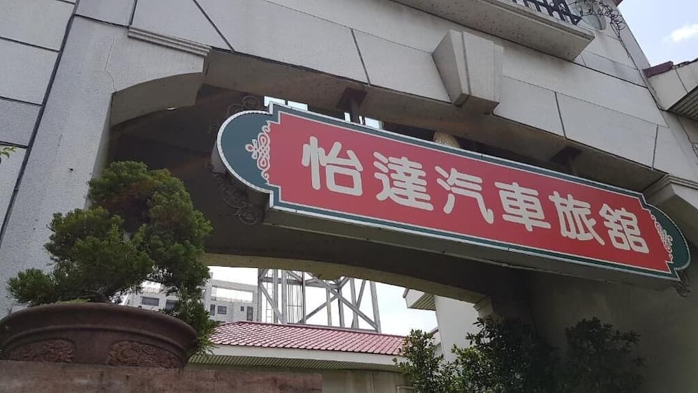 A vibrant red sign reading "怡達汽車旅館" adorns an urban building entrance, framed by lush greenery and architectural details, ideal for travelers seeking accommodation in a bustling cityscape.