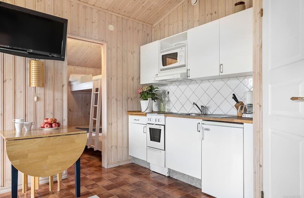 A cozy cabin interior features a compact kitchen with white appliances, wooden walls, a loft bed accessed by a ladder, and a dining table set with apples, ideal for a serene getaway or vacation rental.